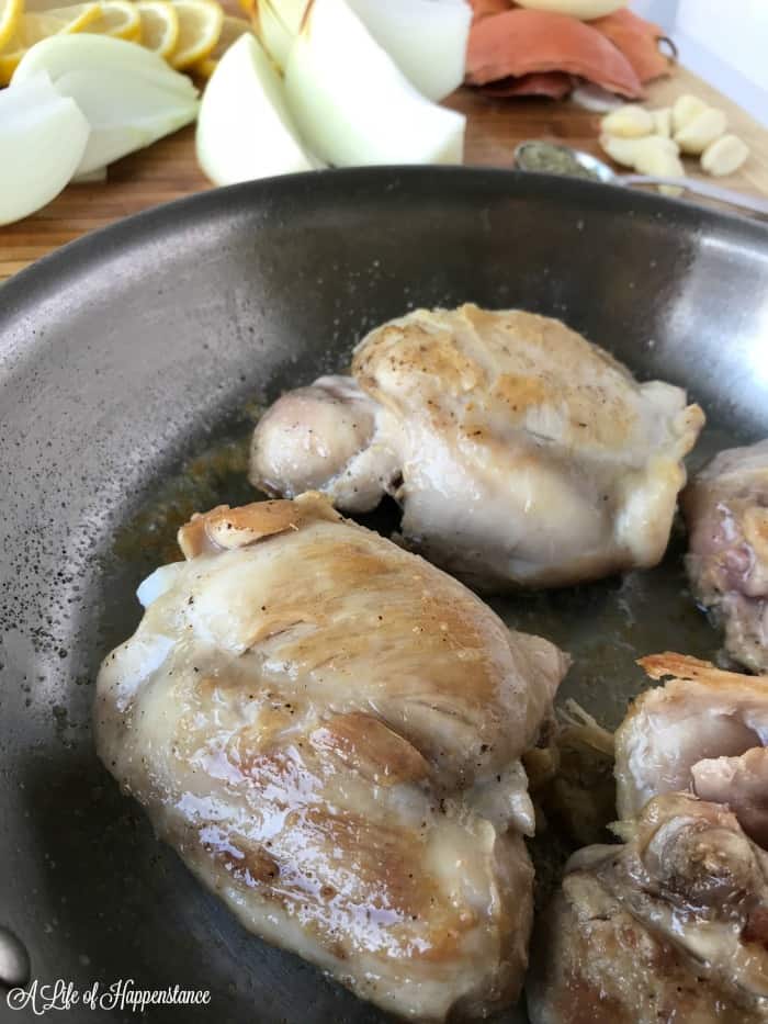 Seared chicken thighs in a skillet with onion wedges and lemon slices in the background. 