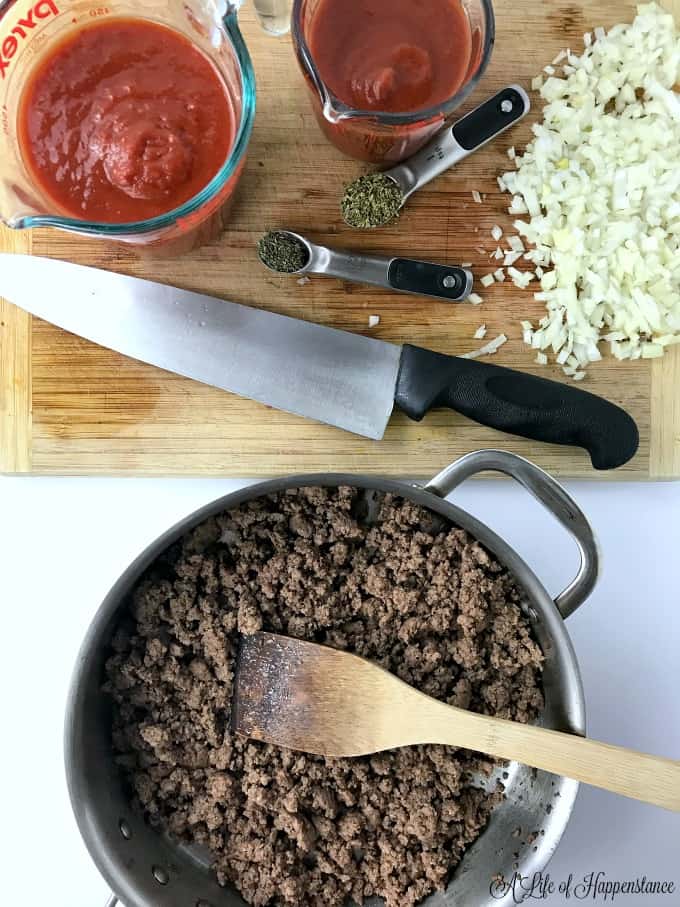Browned ground beef in a saucepan. Onions, tomatoes, herbs on a wooden cutting board. 