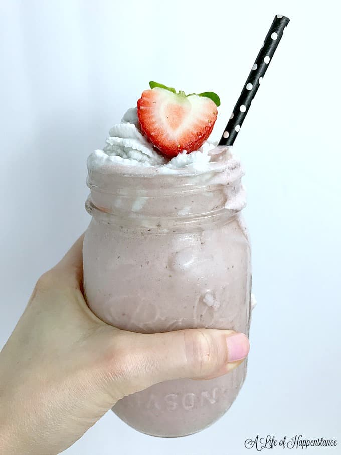 A hand holding a mason jar filled with the smoothie and garnished with coconut whipped cream and a strawberry. 