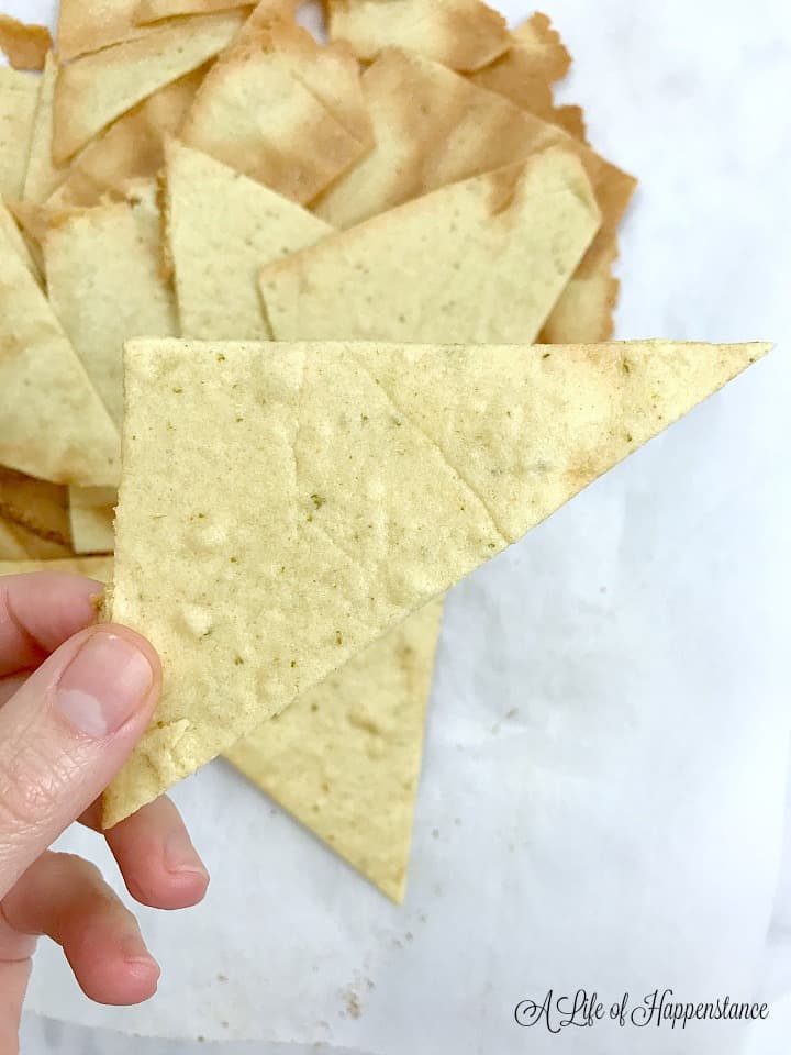 A hand holding a cumin lime tortilla chip with the rest of the chips in the background. 