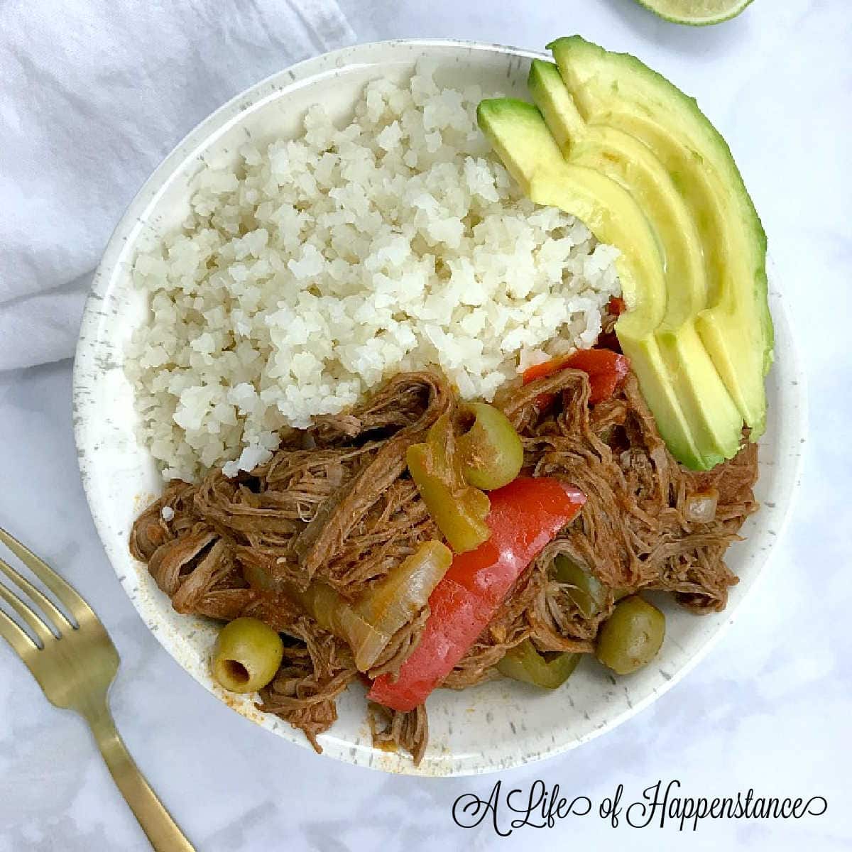 A white bowl filled with Cuban ropa vieja, cauliflower rice, and sliced avocado.