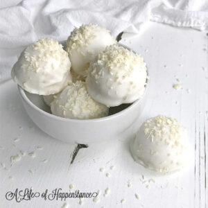 Coconut truffles in a white bowl with one on the table in front of the bowl.