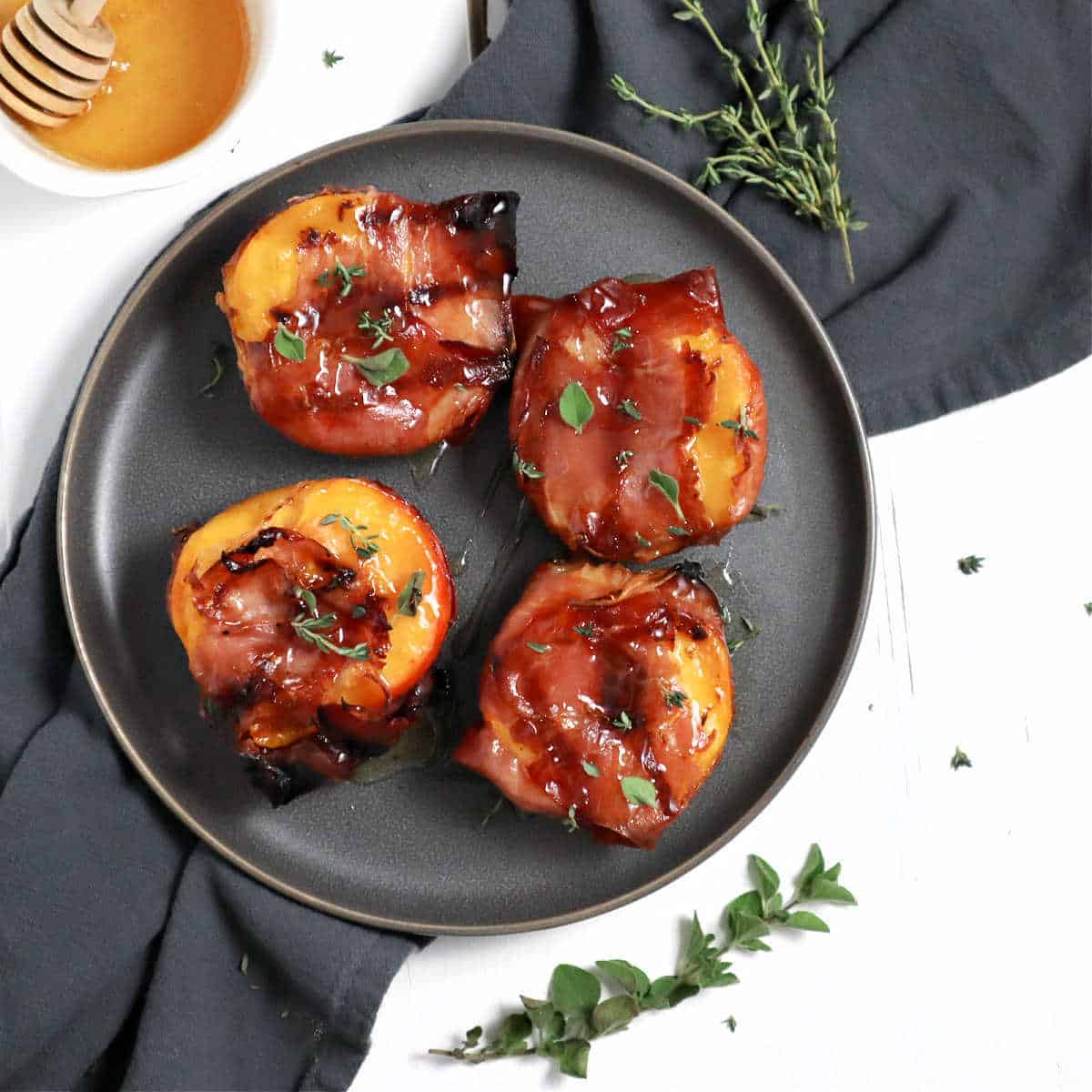 The grilled peaches on a plate surrounded by honey and fresh herbs.