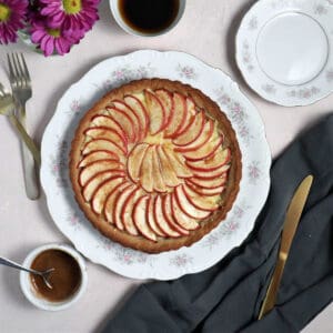 An apple tart on a plate surrounded by utensils, an a vase of flowers.