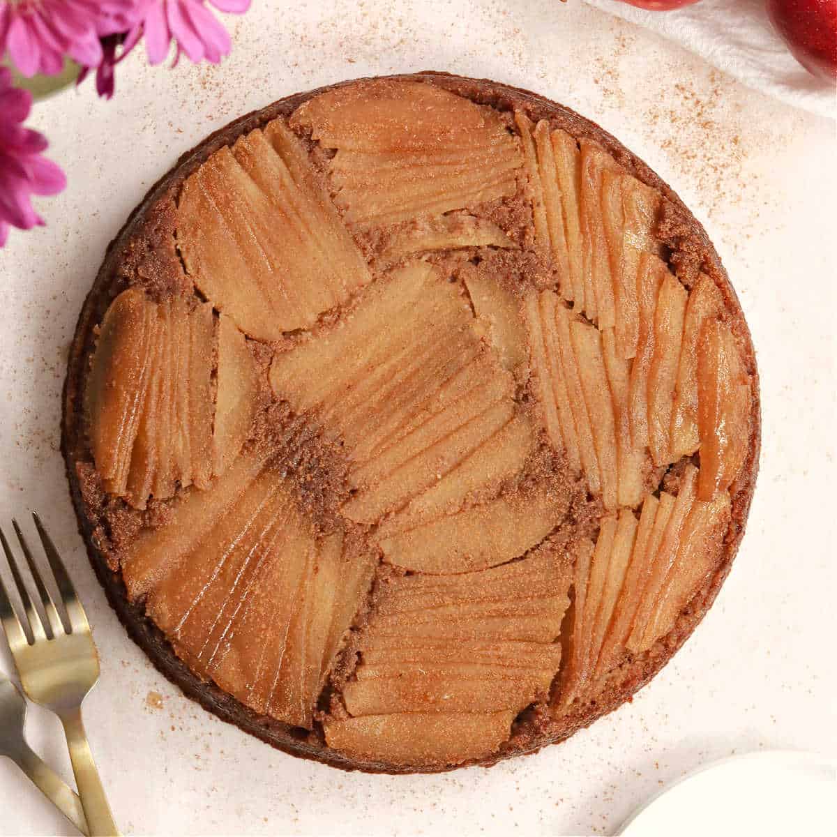 A paleo apple cake on a table surrounded by a couple forks, flowers, and apples.